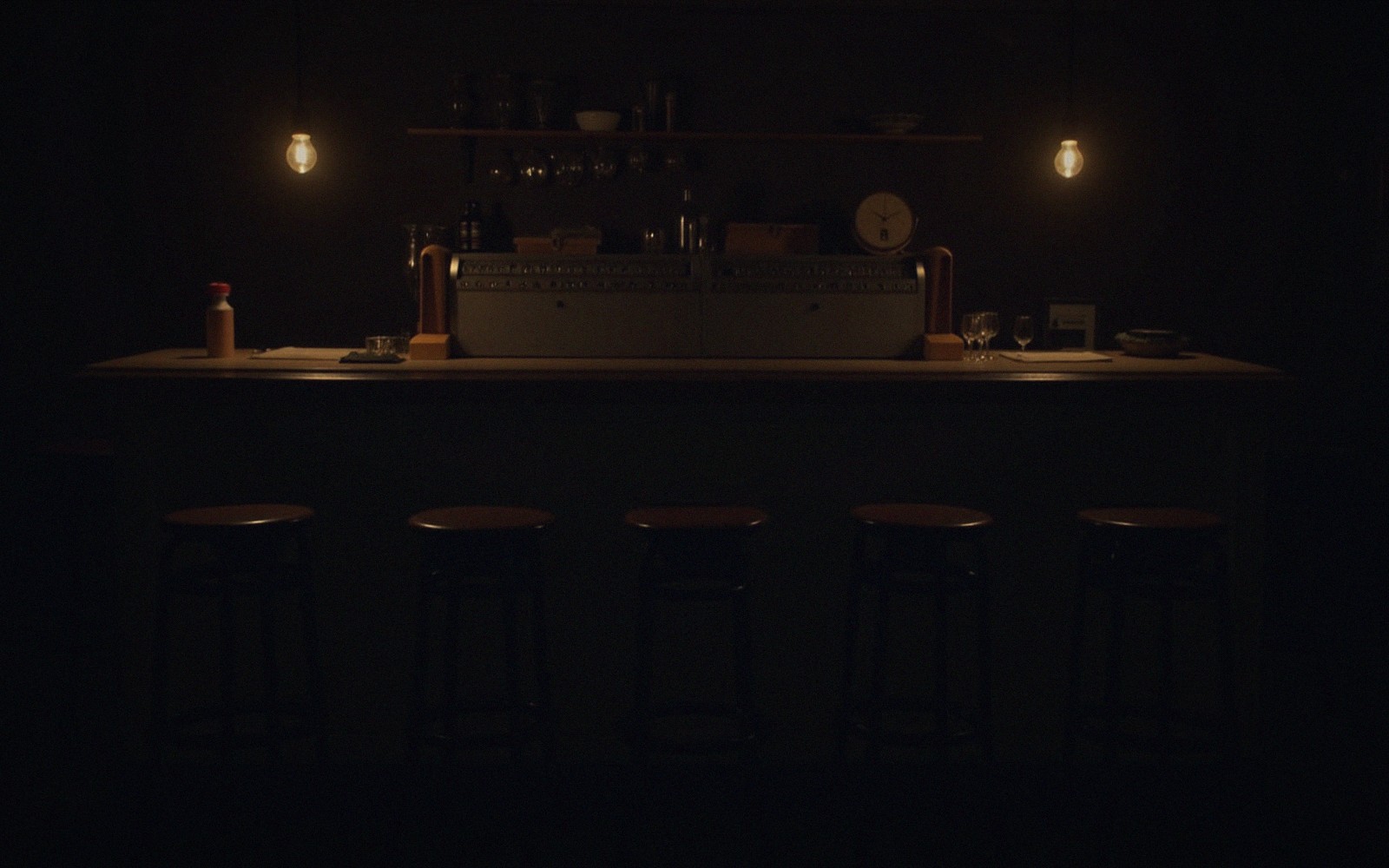 Empty stools line a dimly lit bar counter beneath two bare hanging bulbs